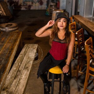 May include: A woman wearing a red and black top with a black hat sits on a stool in a bar setting. The stool is brown and has a black seat. The woman is looking at the camera.