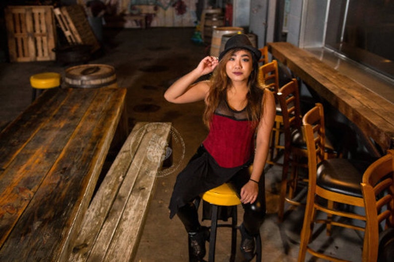 May include: A woman wearing a red and black top with a black hat sits on a stool in a bar setting. The stool is brown and has a black seat. The woman is looking at the camera.