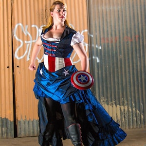 May include: A woman wearing a blue and red corset with a white lace trim and a blue skirt with a red, white, and blue star pattern. She is holding a red, white, and blue shield with a white star in the center. The woman is standing in front of a rusty metal wall.