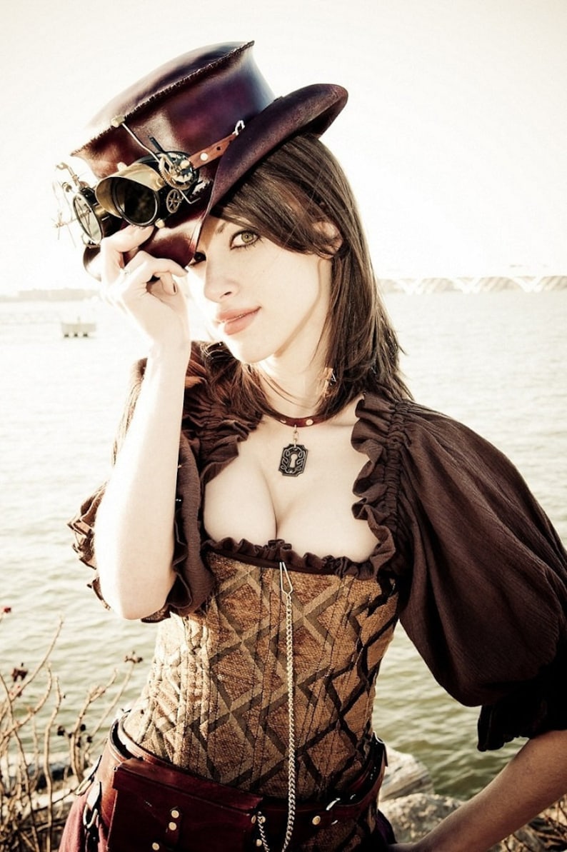 May include: A woman wearing a brown and black steampunk-style hat with goggles, a brown top with a ruffled neckline, a brown corset with a gold chain, and a brown skirt with a belt.