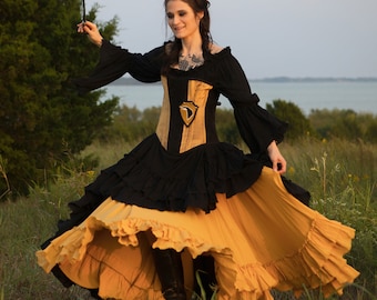 May include: A black and gold Renaissance-style dress with a ruffled skirt and long sleeves. The bodice is black and gold with a decorative element. The dress is worn outdoors in a grassy field.