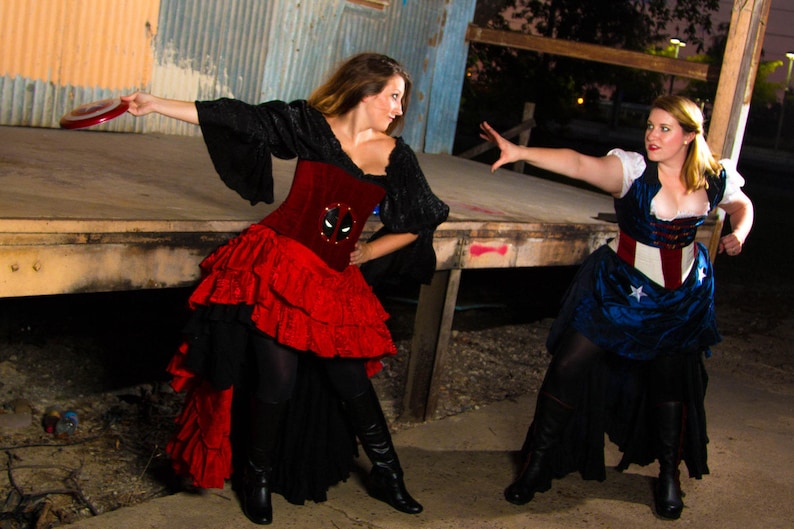 May include: Two women dressed in superhero-themed costumes. One woman is wearing a red and black costume with a Deadpool logo on the bodice. The other woman is wearing a blue and red costume with a red, white, and blue star-spangled belt. Both women are posing dramatically.