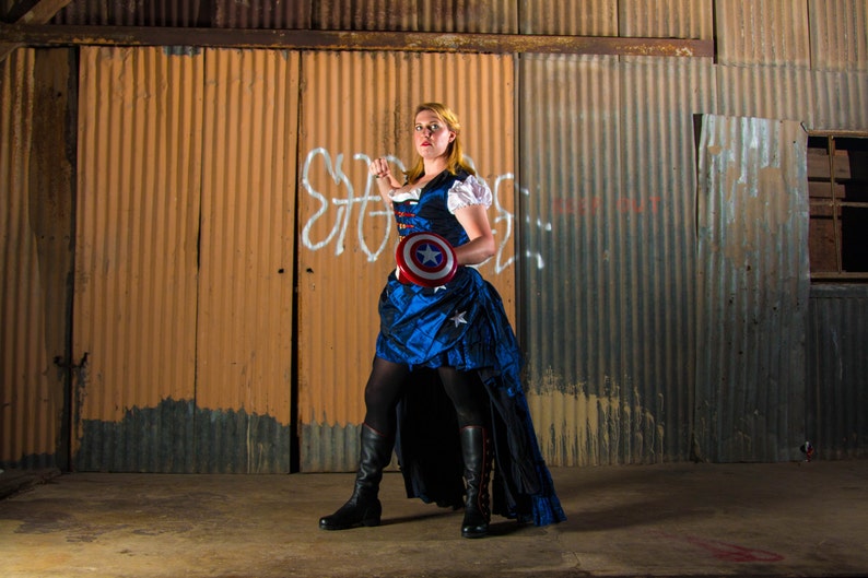 Op de afbeelding: Een vrouw in een blauwe en witte jurk met een rood, wit en blauw sterrendesign houdt een rood, wit en blauw schild vast met een ster in het midden. Ze staat voor een gegolfde metalen muur.