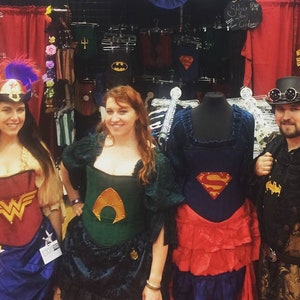 Puede incluir: Tres personas con cors&eacute;s tem&aacute;ticos de superh&eacute;roes, con una cuarta persona con un atuendo de estilo steampunk. Los cors&eacute;s son rojo, verde y azul, con el logotipo de Superman en el cors&eacute; azul. El atuendo steampunk es negro y dorado con un sombrero de copa y gafas.
