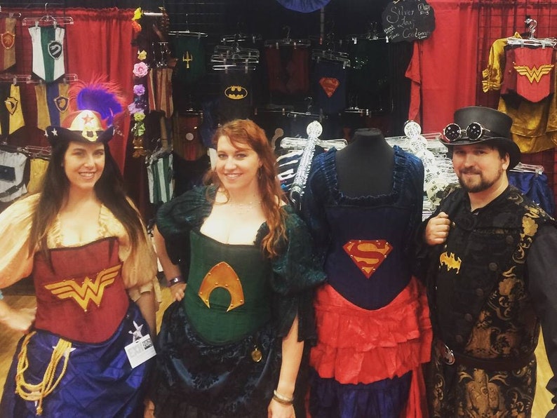Puede incluir: Tres personas con cors&eacute;s tem&aacute;ticos de superh&eacute;roes, con una cuarta persona con un atuendo de estilo steampunk. Los cors&eacute;s son rojo, verde y azul, con el logotipo de Superman en el cors&eacute; azul. El atuendo steampunk es negro y dorado con un sombrero de copa y gafas.