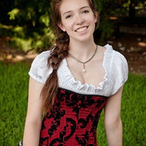 May include: A woman wearing a red and black corset with a white blouse and a long braid. She is sitting on a grassy surface with a green background.