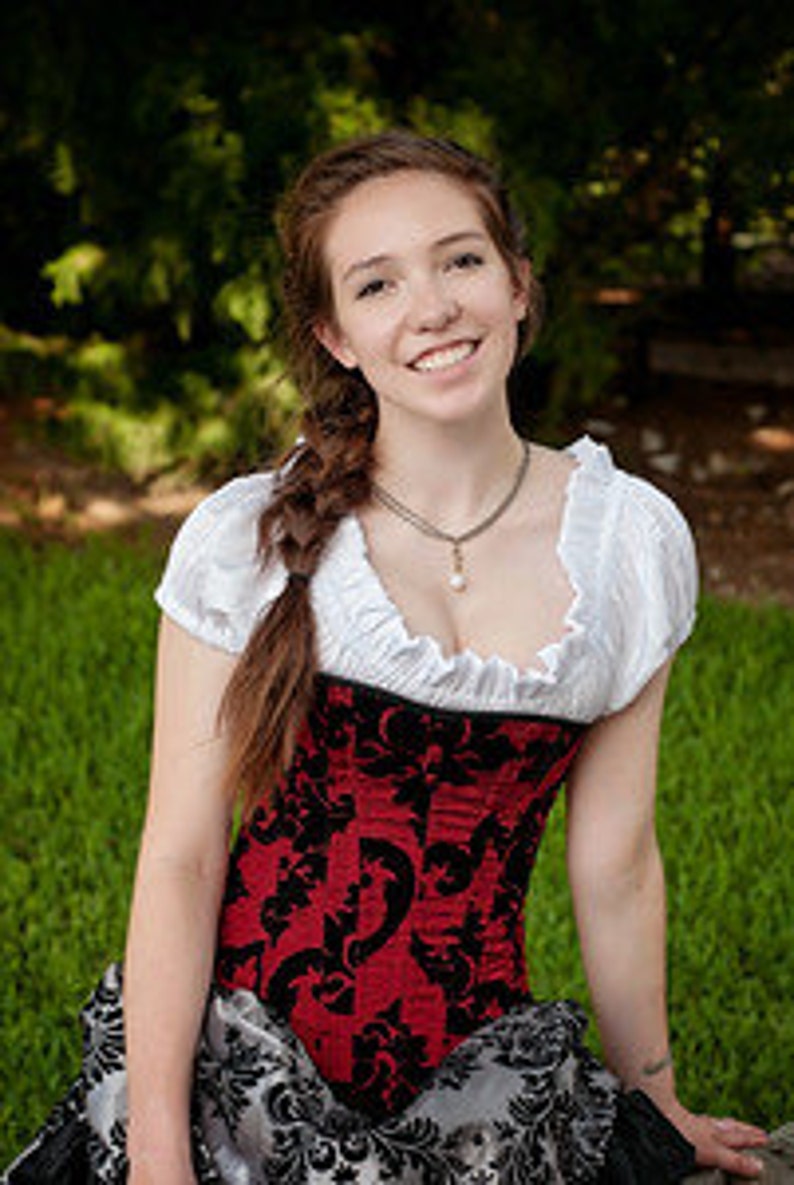 May include: A woman wearing a red and black corset with a white blouse and a long braid. She is sitting on a grassy surface with a green background.