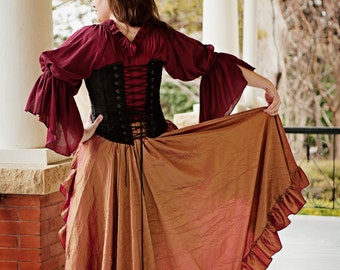 May include: A woman wearing a burgundy top with a black corset and a long, rust-colored skirt with ruffles. The skirt is flowing and the woman is standing on a porch.