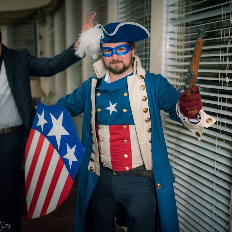 May include: A person dressed as a historical figure wearing a blue and white costume with a red, white and blue shield and a blue and white hat. The person is holding a small, black gun in their right hand.