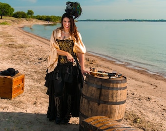 Könnte beinhalten: Eine Frau in einem gold-schwarzen Piratenkostüm steht an einem Sandstrand mit einem Holzfass, einer Truhe und einer Pistole. Sie trägt einen schwarzen Hut mit Pfauenfedern.