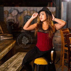 May include: A woman wearing a red corset with black mesh detailing and a black skirt. She is wearing a black fedora hat and has long brown hair. She is sitting on a bar stool in a dimly lit bar setting.