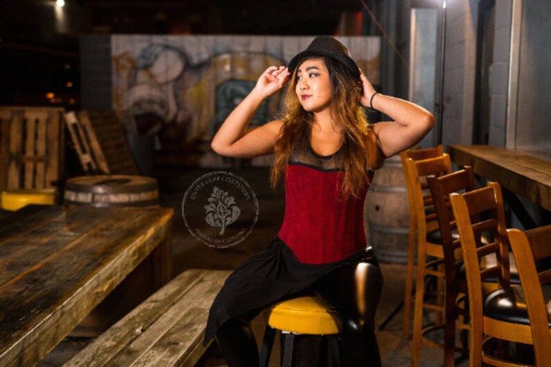 May include: A woman wearing a red corset with black mesh detailing and a black skirt. She is wearing a black fedora hat and has long brown hair. She is sitting on a bar stool in a dimly lit bar setting.
