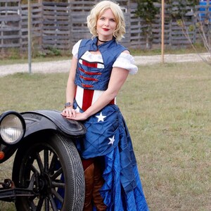 May include: A woman wearing a blue and red corset with a white star pattern skirt, standing next to a vintage car. She is wearing brown boots and has blonde hair.
