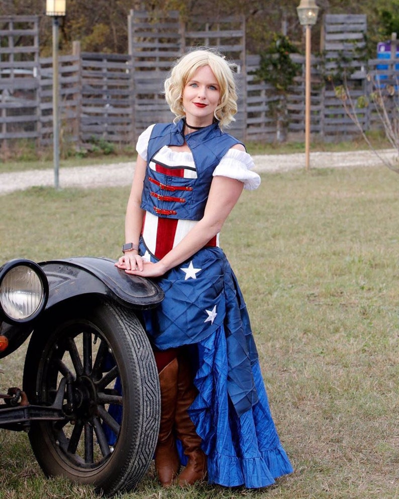 May include: A woman wearing a blue and red corset with a white star pattern skirt, standing next to a vintage car. She is wearing brown boots and has blonde hair.