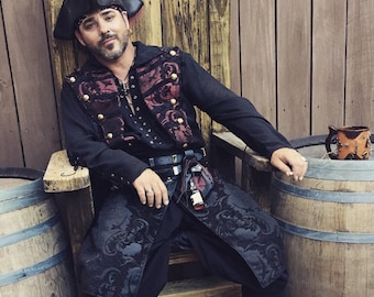 May include: A man wearing a black pirate costume with a red and black patterned vest, a black hat with a skull and crossbones, and a black belt with a silver buckle. He is sitting on a wooden chair with a barrel behind him.