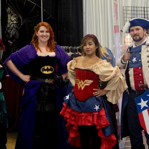 May include: Four people dressed in costumes. A woman in a purple and black dress with a Batman logo, a woman in a red and blue dress with a Wonder Woman logo, a woman in a green dress, and a man in a blue and red costume with a Captain America shield.