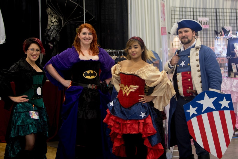 May include: Four people dressed in costumes. A woman in a purple and black dress with a Batman logo, a woman in a red and blue dress with a Wonder Woman logo, a woman in a green dress, and a man in a blue and red costume with a Captain America shield.