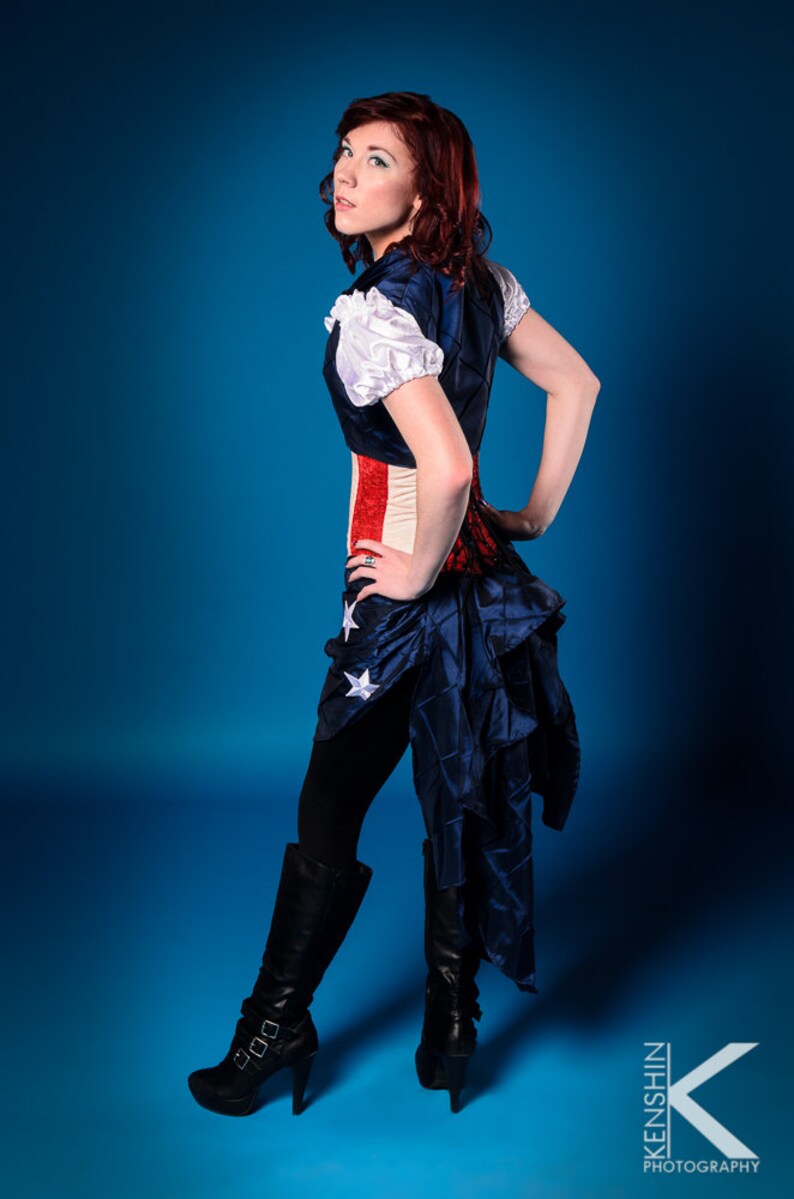 May include: A woman wearing a blue and red corset with white stars and a blue skirt with white stars. She is wearing black tights and black boots with buckles. The background is blue.