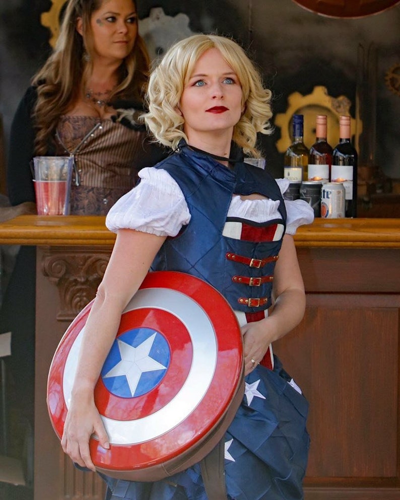 May include: A woman wearing a blue and red vest with a white shirt and a blue and white skirt holds a red, white and blue Captain America shield.