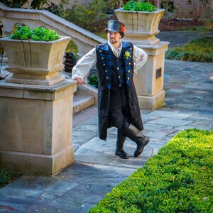 May include: A man wearing a black top hat, a white shirt, a blue vest, and a black jacket with gold trim stands on a stone patio. He is wearing black boots and has a yellow flower pinned to his jacket. The man is standing in front of a stone planter with green plants.