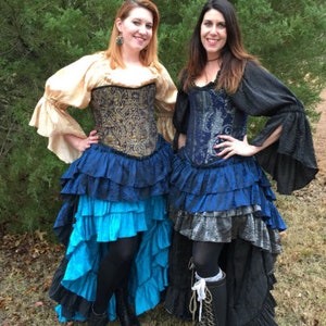 May include: Two women wearing blue and gold corsets with layered ruffle skirts. The skirts are black, blue, and silver. The women are standing in front of a green bush.