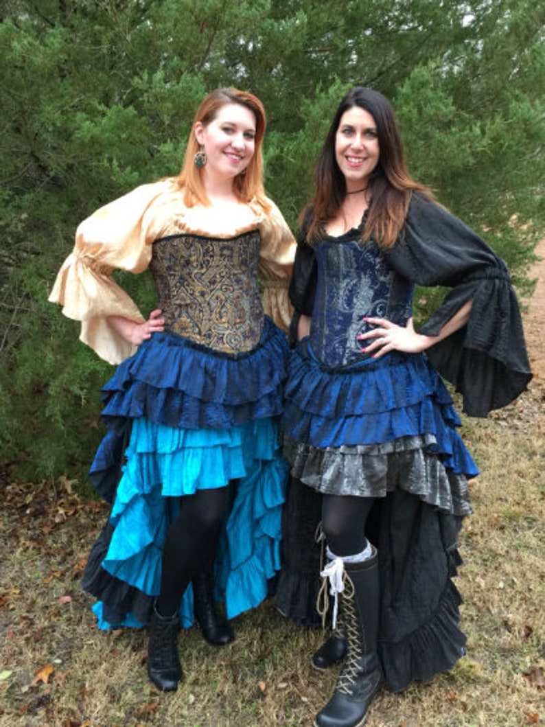 May include: Two women wearing blue and gold corsets with layered ruffle skirts. The skirts are black, blue, and silver. The women are standing in front of a green bush.