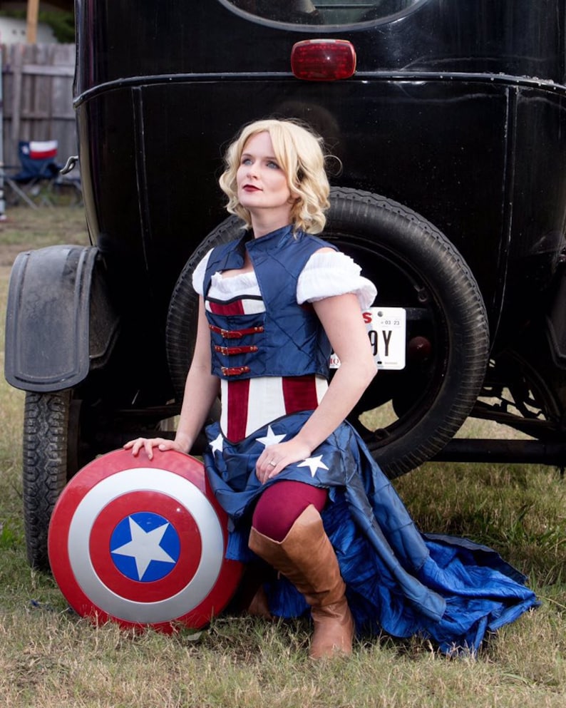May include: A woman dressed in a red, white, and blue costume with a blue and white striped corset, a blue skirt, and red tights. She is holding a Captain America shield in her left hand and is kneeling in front of a vintage car.