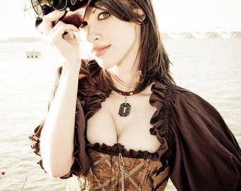 Könnte beinhalten: Eine Frau trägt einen braunen Lederhut mit Steampunk-Brille, eine braune Rüschenbluse, ein braunes und goldenes Korsett und einen braunen Ledergürtel mit goldenen Akzenten. Sie trägt eine Halskette mit einem schlüsselförmigen Anhänger.