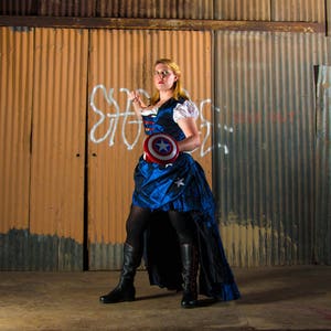 May include: A woman in a blue and white dress holds a red, white, and blue shield with a star in the center. She is standing in front of a corrugated metal wall.