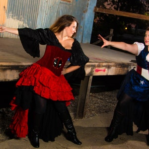 Op de afbeelding: Twee vrouwen gekleed in superhelden kostuums. Een vrouw draagt een rood en zwart kostuum met een Deadpool-maskerontwerp. De andere vrouw draagt een blauw en wit kostuum met een sterrenpatroon en een rood, wit en blauw korset.