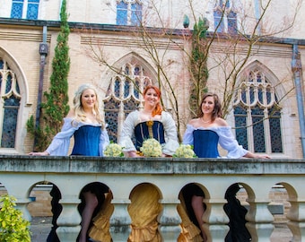 May include: Three women wearing blue and white dresses with off-the-shoulder necklines and white lace sleeves pose in front of a stone building with arched windows. They are holding bouquets of white flowers.