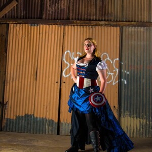May include: A woman wearing a blue and red corset with a white star on it, a blue skirt with a red star on it, and black boots. She is holding a red, white, and blue shield with a white star on it. The background is a rusty metal wall.