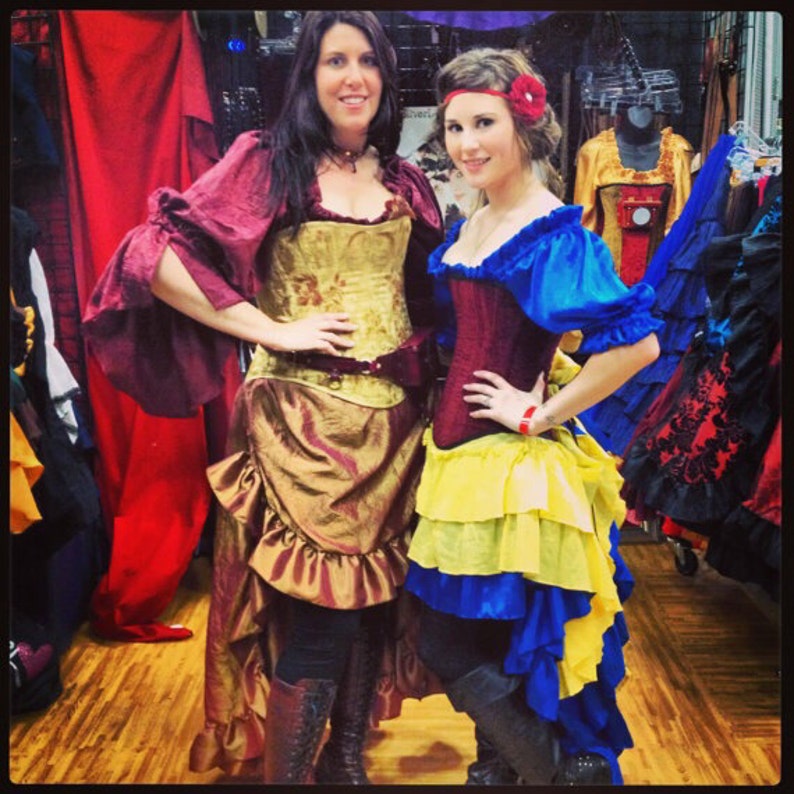 May include: Two women wearing elaborate costumes. One woman is wearing a burgundy and gold corset with a brown skirt and a red jacket. The other woman is wearing a red corset with a yellow and blue skirt and a blue jacket. Both women are smiling.