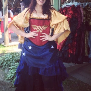 May include: A woman wearing a red and gold corset with a Wonder Woman logo, a blue and white skirt with stars, and a gold shawl.