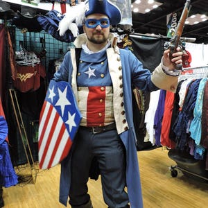 May include: A person dressed as Captain America, wearing a blue and red costume with a white coat, a red, white and blue shield, and a black eye mask. The person is holding a black and silver pistol.