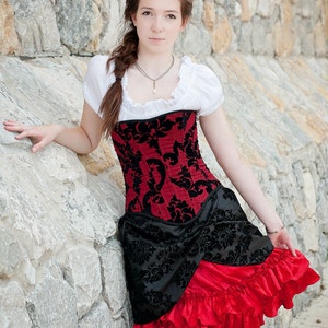 May include: A woman wearing a red and black corset with a white blouse and a red ruffled skirt. She is standing in front of a stone wall.