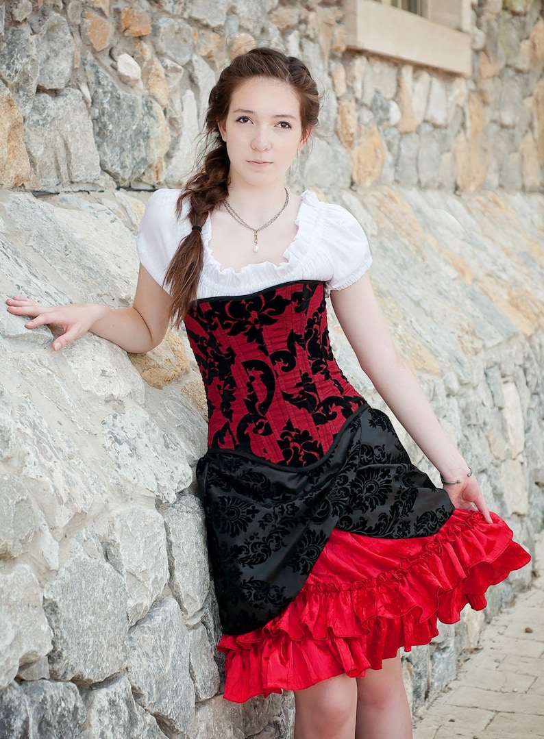 May include: A woman wearing a red and black corset with a white blouse and a red ruffled skirt. She is standing in front of a stone wall.