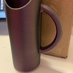 Könnte beinhalten: Eine dunkelviolette Plastiktasse mit Henkel. Die Tasse hat eine breite Öffnung und einen schmalen Boden.