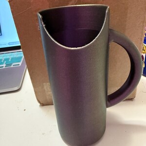 May include: A dark grey ceramic mug with a unique curved handle. The mug has a wide opening and a smooth, matte finish.