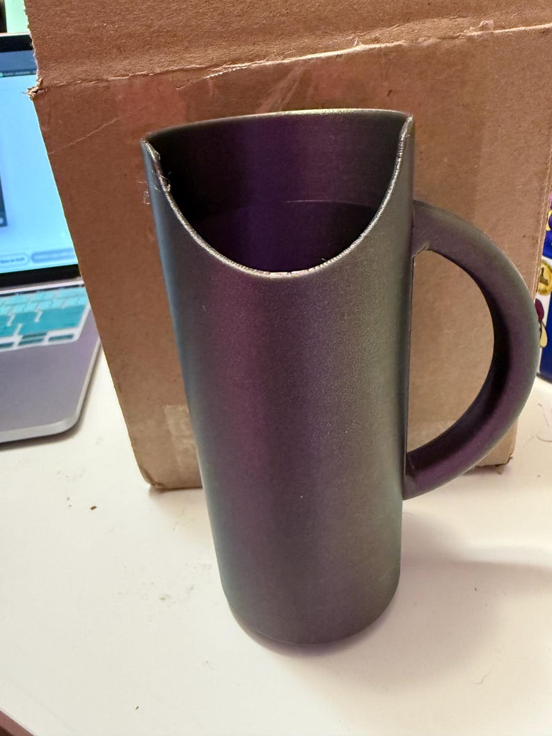 May include: A dark grey ceramic mug with a unique curved handle. The mug has a wide opening and a smooth, matte finish.