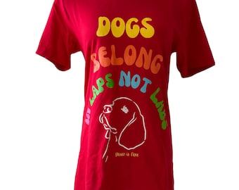 May include: A red t-shirt featuring the slogan "DOGS BELONG IN LAPS NOT LABS" in rainbow-colored text. Below the text is a white outline of a dog's head, with the words "grace is free" beneath it.