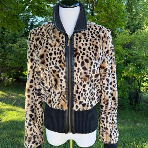 May include: A cropped jacket featuring a tan and black leopard print faux fur body, a black ribbed knit collar, black ribbed knit cuffs, and a black ribbed knit hem. The jacket includes a central vertical black zipper closure with a gold-tone pull.