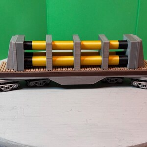 May include: A model train flatcar with a gray frame and wooden deck. It carries two yellow and black cylindrical objects, secured by gray supports. The train car has black wheels and is set against a green backdrop.