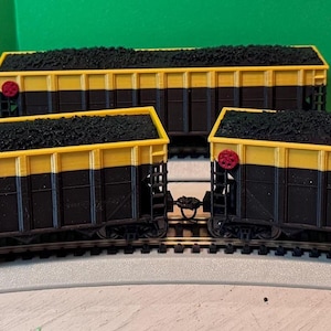 May include: A collection of model train coal cars. Each car is black with a yellow top and filled with black coal. The cars are arranged on a curved section of model train tracks, with a green background.