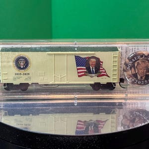 May include: A model train car with a cream-colored body, featuring the Presidential Seal and the years 2025-2029. It also displays an image of a person with an American flag. A round pin with the person's image and the year 2025 is also visible.