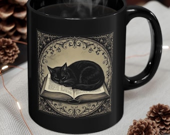 May include: Black ceramic mug featuring a sleeping black cat on an open book, surrounded by an ornate frame. The mug has a black handle and a steam rising from the top, suggesting a hot beverage. Decorative elements include pinecones.
