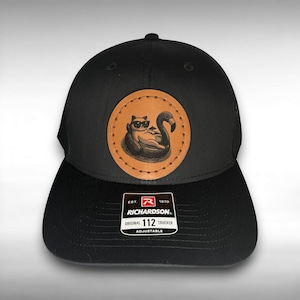 May include: Black trucker hat with a circular leather patch featuring a cat wearing sunglasses lounging on a flamingo float. The patch is stitched onto the hat. The hat has a curved brim and an adjustable snap closure. The Richardson brand label is visible.