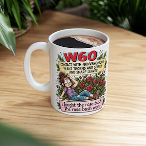 Peut inclure: Un mug en céramique blanche illustré d'un dessin animé représentant une personne assise dans un rosier. Le texte indique W60, CONTACT WITH NONVENOMOUS PLANT THORNS AND SPINES AND SHARP LEAVES, et I fought the rose bush. The rose bush won.