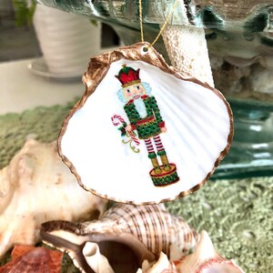 May include: A Christmas ornament made from a seashell, featuring a painted nutcracker design. The shell has a gold-painted edge and a gold string for hanging. The nutcracker is dressed in green and red, holding a candy cane.