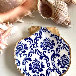 May include: A decorative seashell dish with a blue and white floral pattern. The rim is painted gold. Other seashells are in the background, creating a coastal theme. The dish is ideal for holding small items.